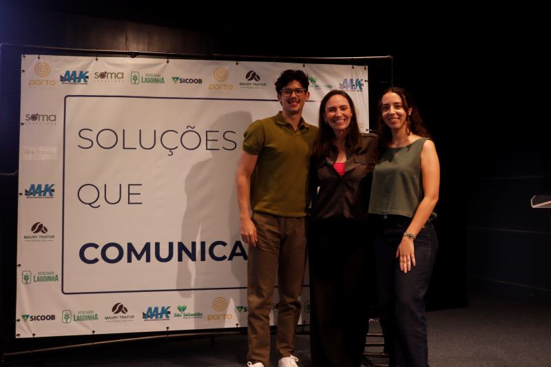 A jornalista Cecília Flesch ao lado de dois membros da PCJ, em frente ao painel do evento Soluções que Comunicam.